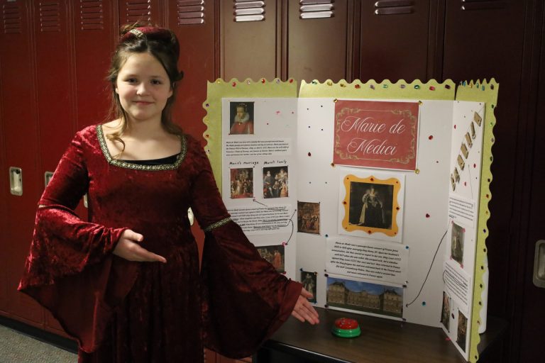Young girl in Renaissance gown presents Marie de Medici project at school display.