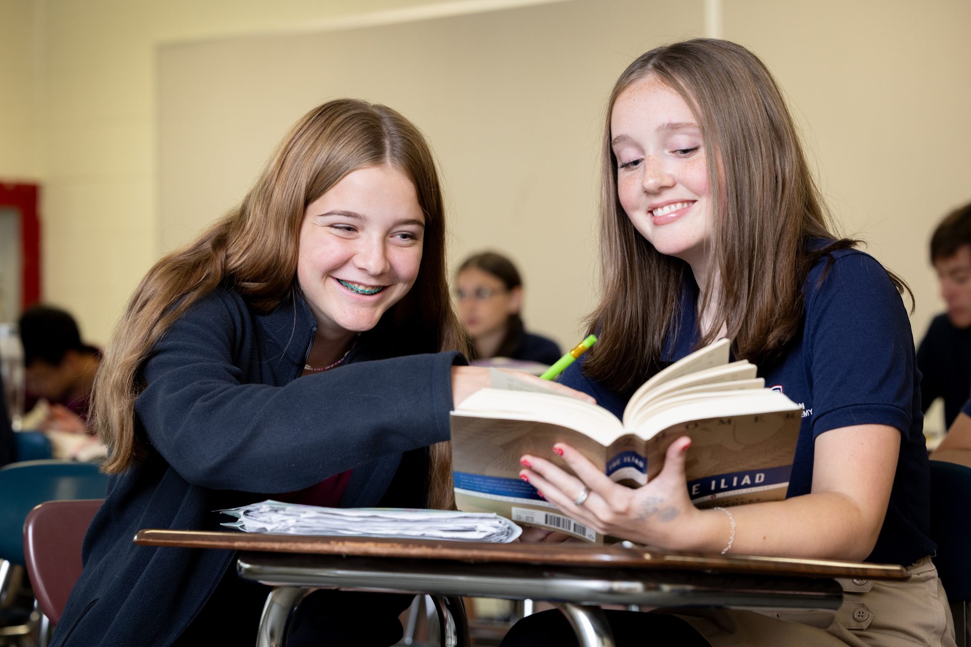 Two girls happily study together over The Iliad in a bright classroom.