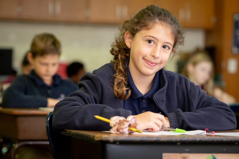 Cheerful girl engaged in classroom learning, writing with a pencil, surrounded by colorful school supplies.