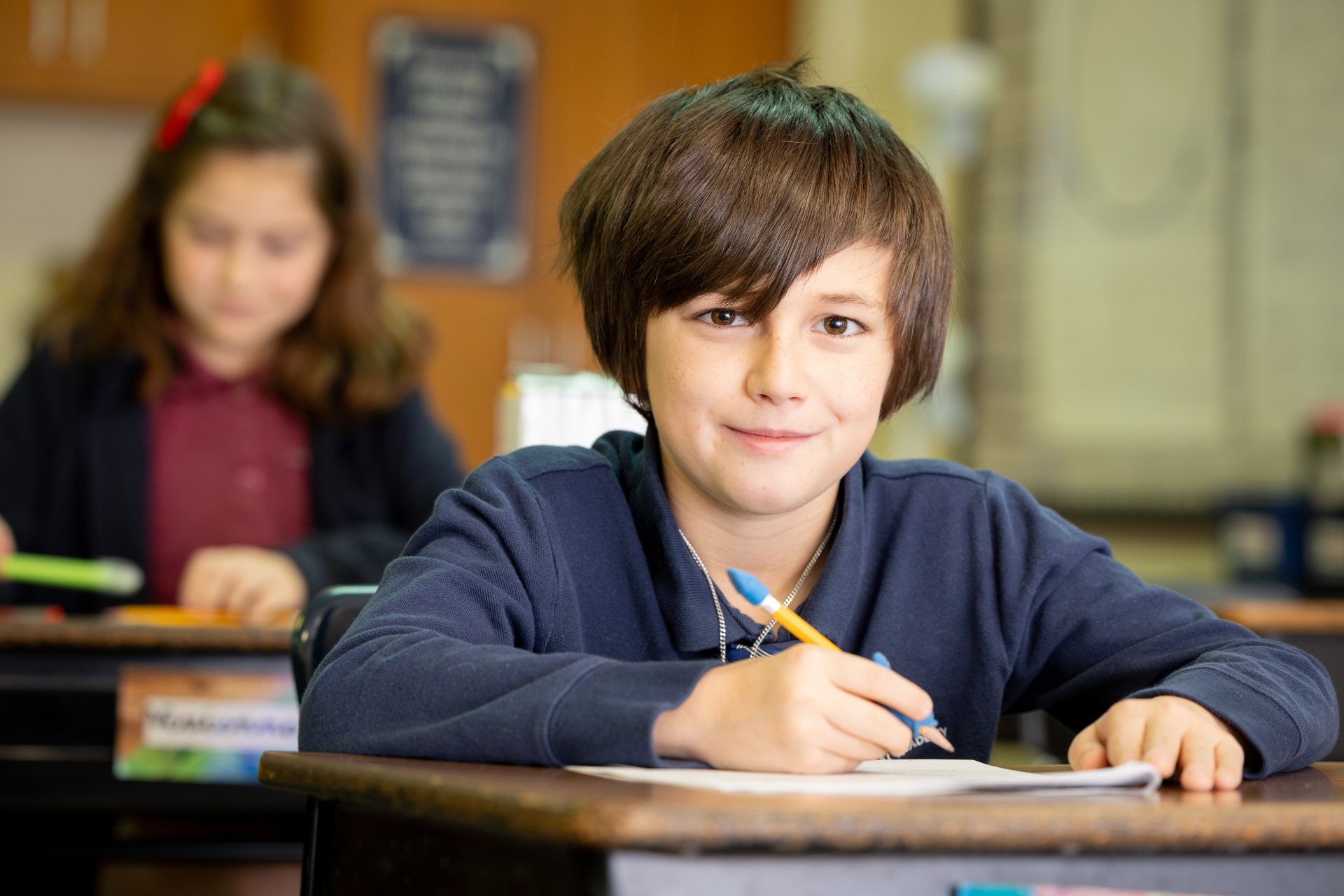 Young boy in navy uniform smiles while writing in a bright classroom, fostering learning.